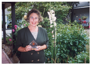 COL photo of a woman standing in a garden holding a small trophy. She is smiling. There are many colourful flowers in bloom in the garden, which is surrounded on three sides by weatherboard buildings.
