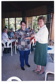 COL photo of a woman presenting an engraved silver platter to another woman, while others (seated at tables), look on.