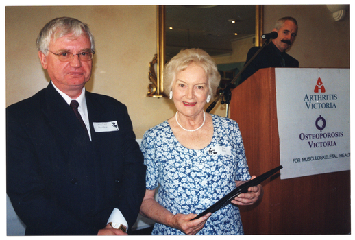 COL photo of a man and a woman standing close to one another. The woman is holding a framed award. Behind them, there is a man standing behind a podium. On the wall behind the podium, there is a large mirror with an ornate gold frame.