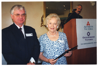 COL photo of a man and a woman standing close to one another. The woman is holding a framed award. Behind them, there is a man standing behind a podium. On the wall behind the podium, there is a large mirror with an ornate gold frame.