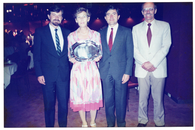 COL photo of four people standing in a row in a large hall. One of them, a woman, is holding a silver platter. In the background, there are several people either seated at tables or standing.
