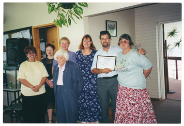 COL photo of a small group of people posing for a photo inside a building. One of them is holding a framed award certificate. Along the wall behind them (to the left of the frame), there is a large PA speaker sitting on a chair which has been placed on a small table. The speaker sits in front of a large noticeboard on the wall. Next to the wall, there is an doorway, with the door ajar. On the right side of the frame, a ramp with a wooden ballustrade is visible through an open doorway. Parts of a tall plant (partially obscured) is also visible. There is another smaller plant hanging from the ceiling above where the group is standing.