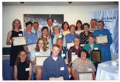 COL photo of a large group of people of varying ages posing for a photo. Many of them are holding up a framed award. On the wall behind them (partially obscured) is a large mural-sized black and white Photograph of an athletics running event. Also behind them, on the left, is a white and blue Move It Or Lose It banner. Beside them is a large round table with a white table cloth & crockery.