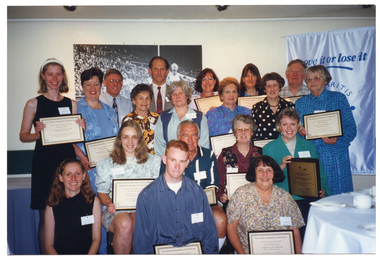 COL photo of a large group of people of varying ages posing for a photo. Many of them are holding up a framed award. On the wall behind them (partially obscured) is a large mural-sized black and white Photograph of an athletics running event. Also behind them, on the left, is a white and blue Move It Or Lose It banner. Beside them is a large round table with a white table cloth & crockery.