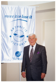 COL photo of a man standing beside a tall freestanding vertical banner. Behind him is a panelled wall. The banner is white with blue text and two logos. The title on the banner is "move it or lose it". Below the title are the text and logos for Arthritis Victoria and VicHealth.