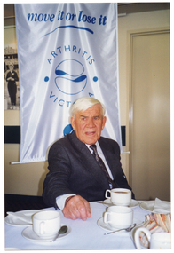 COL photo of a man sitting at a round table covered with a white tablecloth. On the table are cups and saucers, small plates with serviettes, and a plate of sandwiches. Behind him is a tall freestanding vertical banner. The banner is white with blue text and two logos. The title on the banner is "move it or lose it". Below the title are the text and logos for Arthritis Victoria and VicHealth. On the wall behind the banner, there is a large framed black and white photo (partially obscured). The photo depicts some sports persons standing on podiums (partially obscured), with a large crowd of spectators sitting in the grandstand behind them.
