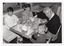 B&W photo of a man and three children. They are sitting at a table on a short bench and two seats. On the table are hamburgers, soft drinks, fries and two printed cardboard headbands.