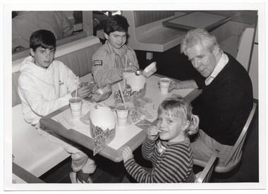 B&W photo of a man and three children. They are sitting at a table on a short bench and two seats. On the table are hamburgers, soft drinks, fries and two printed cardboard headbands.