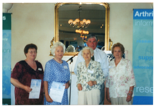 COL blurred photo of a small group of people standing in front of a gold-framed mirror. Two of them are holding up their award certificates. Beside them, to the left, is a blue and green Arthritis Victoria banner.