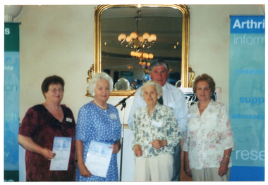 COL blurred photo of a small group of people standing in front of a gold-framed mirror. Two of them are holding up their award certificates. Beside them, to the left, is a blue and green Arthritis Victoria banner.