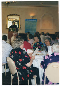 COL photo of groups of people seated at large round tables in a large hall. Some people are looking at a piece of printed paper, which they are holding in their hands. In the background, a man is speaking at a lectern. Behind him is a blue and green Arthritis Victoria banner.