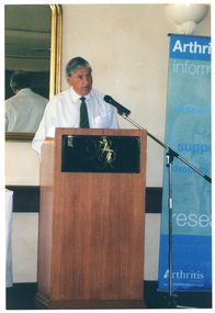 COL photo of a man standing at a wooden lectern, speaking into a microphone. On the front of the lectern is the monogram HR in gold on a black background. Behind him is a large gold-framed mirror and beside him is a blue and green Arthritis Victoria banner.