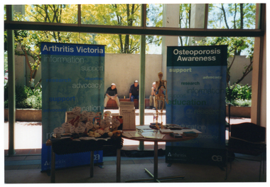COL photo of two small display tables in front of two vertical banners. Behind them is a wall of glass windows. On one of the tables is a range of food, each with a large printed label. On the other table are some brochures and leaflets, and a small skeleton on a stand. Through the window, a few people are sitting on a low tiled ledge outside.