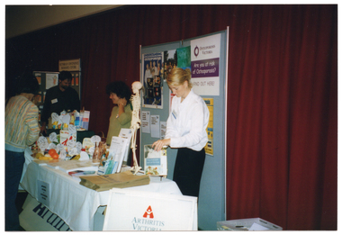 COL photo of a woman browsing the contents of some display tables, with attendants standing behind the tables. One table contains a range of food, each with a large printed label, while the other has some printed material (books and/or brochures) on top. Behind the attendents, there are some display boards containing posters and other information. Along the wall behind them hangs a long red curtain.