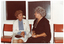 COL photo of two women chatting as they sit on two vinyl chairs in front of a window. There are four orange vinyl chairs in total.