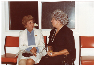 COL photo of two women chatting as they sit on two vinyl chairs in front of a window. There are four orange vinyl chairs in total.