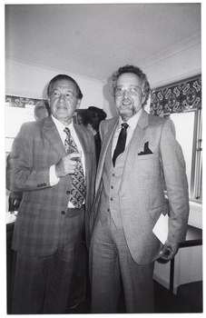 B&W photo of two people standing near the corner of a room with large windows. One of them is holding a drink in his hand. Behind them, there are two other people standing in the corner.