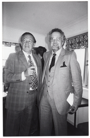 B&W photo of two people standing near the corner of a room with large windows. One of them is holding a drink in his hand. Behind them, there are two other people standing in the corner.