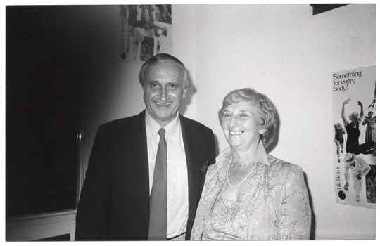 B&W photo of two people standing in the corner of a room, smiling. Behind them is a poster on each wall, depicting people exercising. The poster on the right includes the text "Life Be In It" and "Something for every body!"
