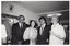 B&W photo of four people standing close together, in a row. One of them is holding a drink in her hand. In the background, there are several people standing beside and behind them.