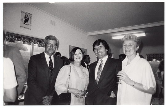 B&W photo of four people standing close together, in a row. One of them is holding a drink in her hand. In the background, there are several people standing beside and behind them.