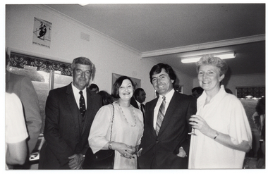 B&W photo of four people standing close together, in a row. One of them is holding a drink in her hand. In the background, there are several people standing beside and behind them.