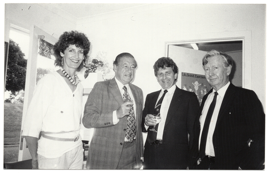 B&W photo of four people standing close together in the corner of a room. The two people in the middle are each holding a drink in their hand. Behind them, there is a large window and an open door. There is a "Life Be In It" exercise promotion poster on the door.