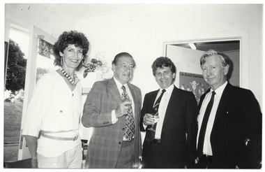 B&W photo of four people standing close together in the corner of a room. The two people in the middle are each holding a drink in their hand. Behind them, there is a large window and an open door. There is a "Life Be In It" exercise promotion poster on the door.