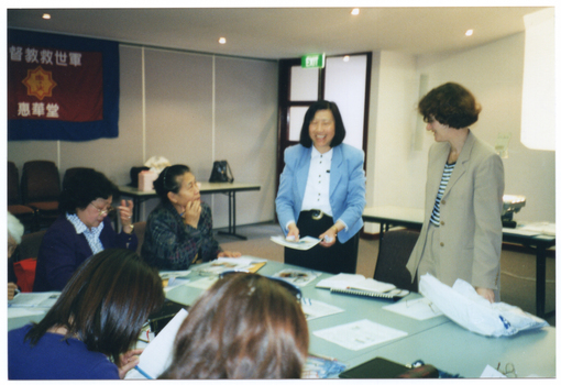 COL photo of two women standing at some tables in a hall, while several other women are seated at the table. One of the women standing is holding some printed material in her hands. Hanging on the wall is a large banner with a heading and sub-heading above and below a logo. The characters are in an Asian language. According to Google Translate, the characters are in the Chinese language. In the heading, they spell out the words "Christian Salvation Army", and "Keikado" in the sub-heading.