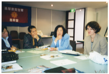 COL photo of several women seated at a group of tables pushed together. On the tables is some printed material, such as brochures, books, and booklets. Hanging on the wall is a large banner with a heading and sub-heading above and below a logo. The characters are in an Asian language. According to Google Translate, the characters are in the Chinese language. In the heading, they spell out the words "Christian Salvation Army", and "Huihuatang" in the sub-heading.