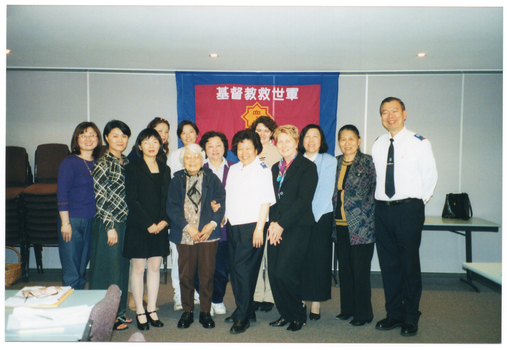 COL photo of a large group of people posing for a photo in a hall. Two people in the group are wearing a uniform with a logo on their shirt epaulettes. Behind the group is a large banner containing a heading in an Asian language and a logo. According to Google Translate, the characters in the heading are in the Chinese language and spell out the words, "Christian Salvation Army".