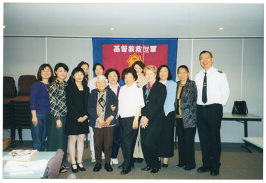COL photo of a large group of people posing for a photo in a hall. Two people in the group are wearing a uniform with a logo on their shirt epaulettes. Behind the group is a large banner containing a heading in an Asian language and a logo. According to Google Translate, the characters in the heading are in the Chinese language and spell out the words, "Christian Salvation Army".