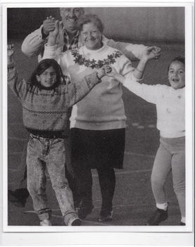 B&W photo of two adults and two children walking hand in hand on an outdoor sports court. They are holding their linked hands up in the air while walking.