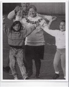 B&W photo of two adults and two children walking hand in hand on an outdoor sports court. They are holding their linked hands up in the air while walking.