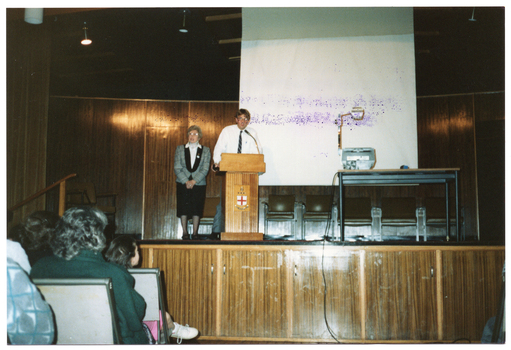 COL photo of a man and a woman standing on a wood-panelled stage, with an audience partially visible in the foreground. The man is speaking at a lectern while the woman stands beside him. Beside them is a table with a slide projector on it, behind which is a large screen. There is some ink contamination on the image surface from a purple ink stamp that was stamped on the verso of another photo.