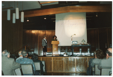 COL photo of two women standing on a wood-panelled stage, presenting to an audience. One woman is speaking at a lectern while the other woman stands beside her. Beside them is a table with a slide projector on it, behind which is a large screen. There is a small degree of ink contamination on the image surface from a purple ink stamp that was stamped on the verso of another photo.