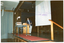 COL photo of a man, standing on a wood-panelled stage. He is facing sideways as he leans into a lectern while presenting to an audience. On a table beside him is a slide projector, and there is a large screen behind him. There is some ink contamination on the image surface from a purple ink stamp that was stamped on the verso of another photo.