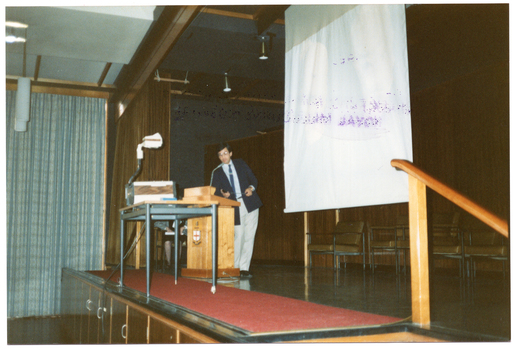 COL photo of a man, standing on a wood-panelled stage. He is facing sideways as he leans into a lectern while presenting to an audience. On a table beside him is a slide projector, and there is a large screen behind him. There is some ink contamination on the image surface from a purple ink stamp that was stamped on the verso of another photo.