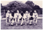 B&W photo of a troupe of dancers in traditional dress. They are standing on the grass, linking hands. Behind them, there is a tall cyclone fence with some large trees or shrubs behind it.