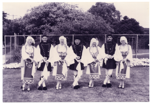 B&W photo of a troupe of dancers in traditional dress. They are standing on the grass, linking hands. Behind them, there is a tall cyclone fence with some large trees or shrubs behind it.