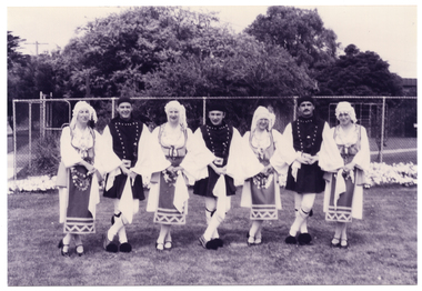 B&W photo of a troupe of dancers in traditional dress. They are standing on the grass, linking hands. Behind them, there is a tall cyclone fence with some large trees or shrubs behind it.