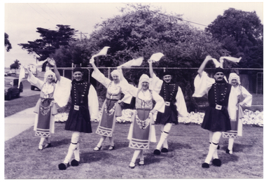 B&W photo of a troupe of dancers in traditional dress. They are standing on the grass in two rows, each holding a handkerchief with one arm held up high. Behind them, there is a tall cyclone fence with some large trees or shrubs behind it.