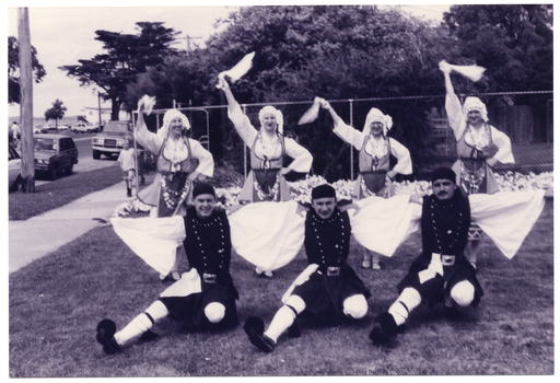 B&W photo of a troupe of dancers in traditional dress. They are on the grass in two rows, holding a dance pose. In the back row, four women standing with one arm raised high and waving a handkerchief. In the front row, three men are kneeling down with one leg outstretched to the front and their arms outstetched to the side, resting on the next dancer's shoulders. Behind them, there is a tall cyclone fence with some large trees or shrubs behind it.