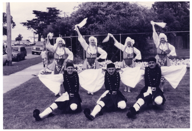 B&W photo of a troupe of dancers in traditional dress. They are on the grass in two rows, holding a dance pose. In the back row, four women standing with one arm raised high and waving a handkerchief. In the front row, three men are kneeling down with one leg outstretched to the front and their arms outstetched to the side, resting on the next dancer's shoulders. Behind them, there is a tall cyclone fence with some large trees or shrubs behind it.