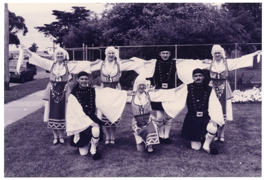 B&W photo of a troupe of dancers in traditional dress. They are on the grass, in two rows: the dancers in the back row are standing, while the dancers in the front row are kneeling on one knee. They are all posing with their arms outstetched, touching the next dancer's shoulders.  Behind them, there is a tall cyclone fence with some large trees or shrubs behind it.
