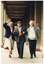 COL photo of three elderly people walking arm in arm along the covered veranda of a large old building.