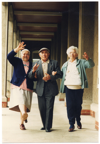 COL photo of three elderly people walking arm in arm along the covered veranda of a large old building.