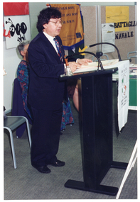 COL photo of a man standing at a black lectern, speaking. There is a small bell placed on the lectern. Partially visible behind and to the side of him is a woman, seated. There are posters on the wall behind them.