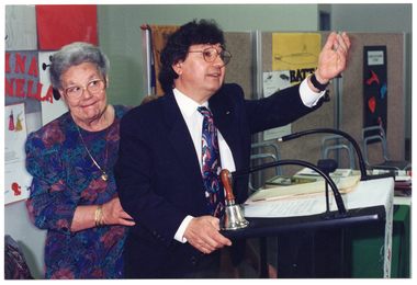 COL photo of a man standing at a black lectern speaking, with one arm outstretched. There is a small bell placed on the lectern. Behind him is a woman with one hand on his waist. There are posters on the wall behind them.