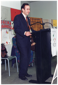 COL photo of a man standing at a black lectern, speaking. There is a small bell placed on the lectern. Partially visible behind and to the side of him are two women, seated. There are posters on the wall behind them.